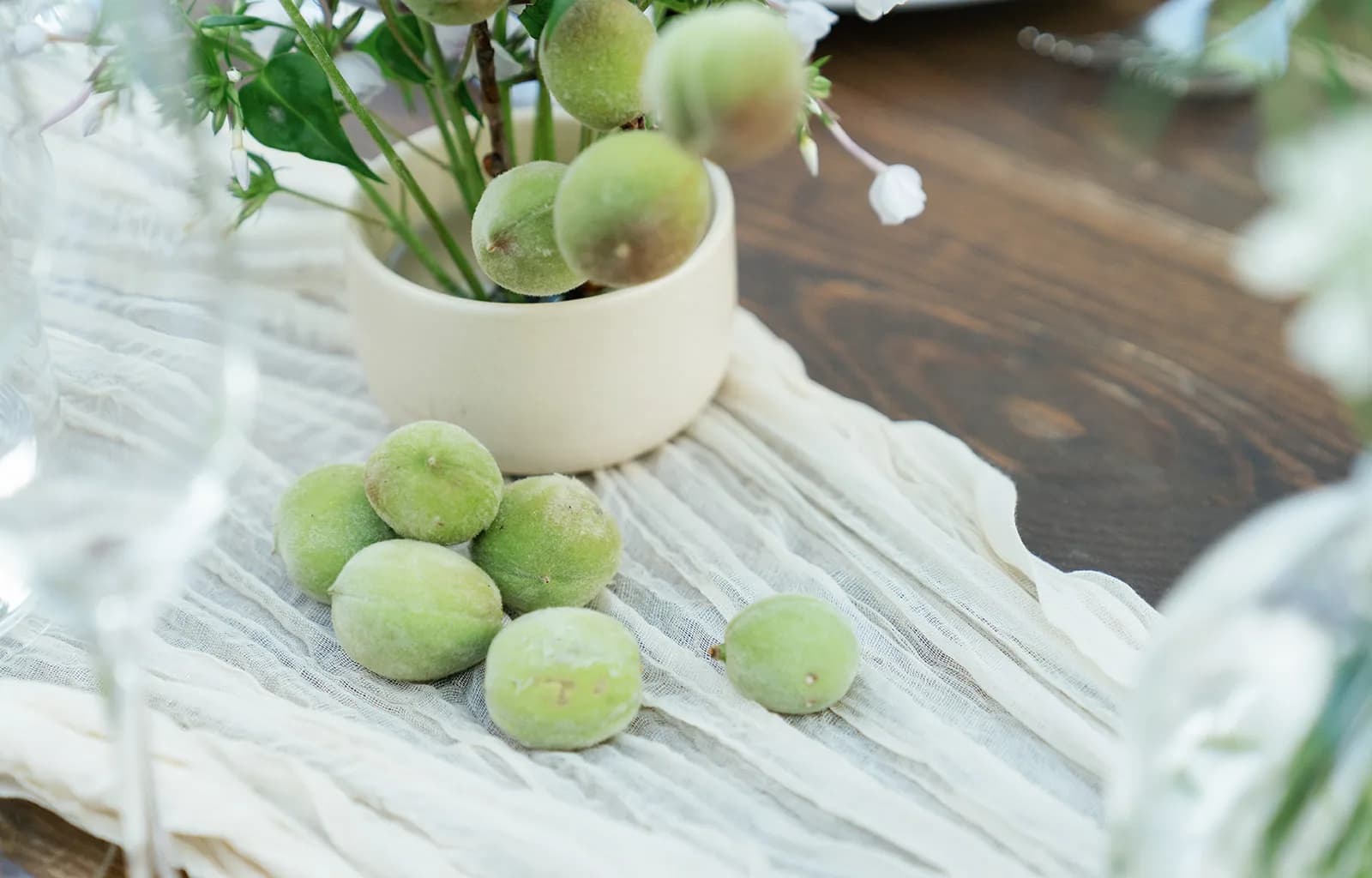 Fuzzy green baby peaches scattered across a gauze runner beside a cream ceramic bowl holding a peach branch and white phlox