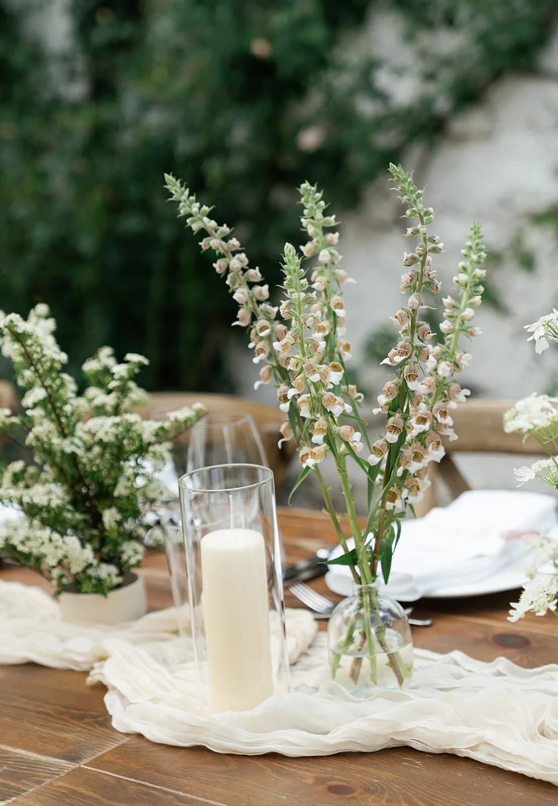 Cream foxglove spires in a clear bud vase beside a pillar candle in glass, spirea in a low cream pot on the gauze runner