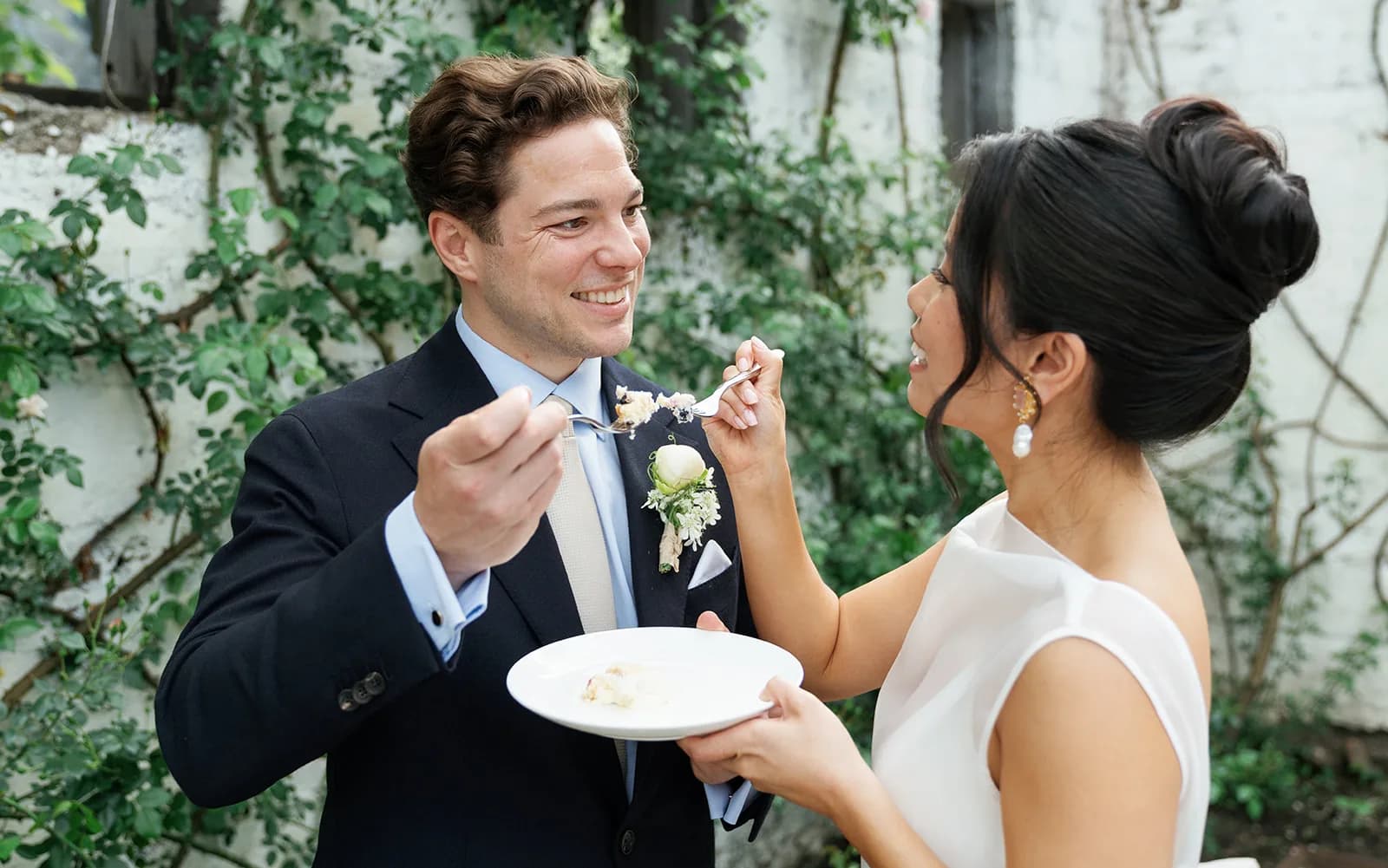 Trisha feeds Jim a bite of cake as he wears a white ranunculus boutonniere, climbing roses on the whitewashed wall behind