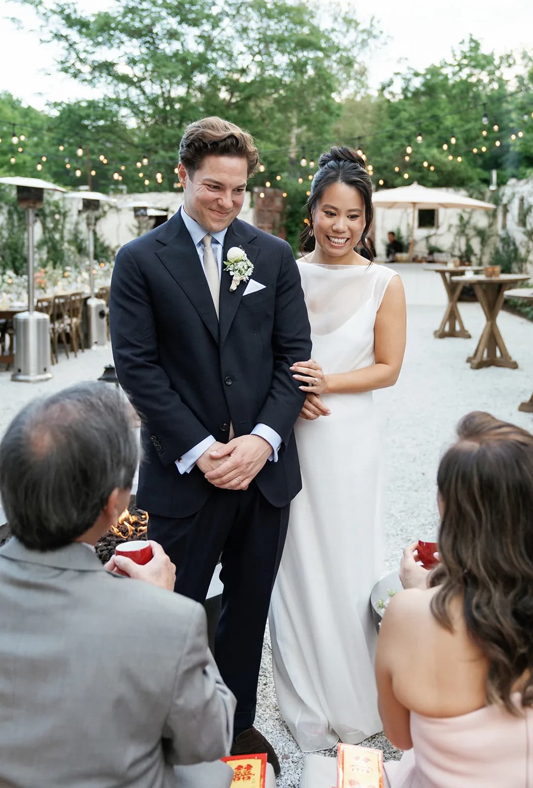 Trisha and Jim stand together during a tea ceremony as seated guests hold red cups, string lights strung over the courtyard