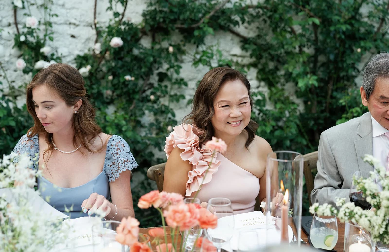 Guests seated along the reception table behind coral ranunculus and white spray blooms, roses climbing the wall behind them