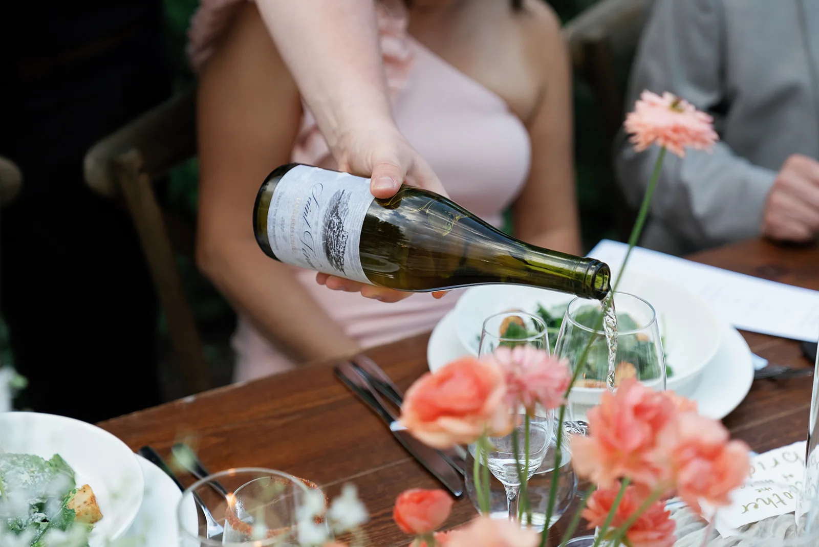 Wine poured into a glass over coral ranunculus and a single coral bloom on a tall stem in the reception tablescape