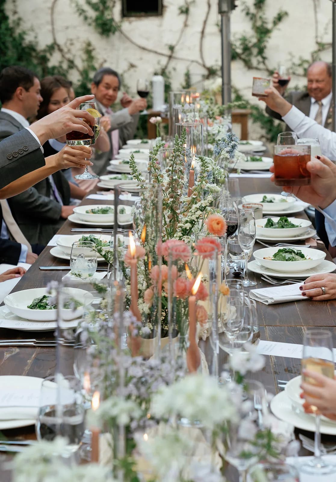 Guests raise glasses in a toast down the length of the table, foxglove, coral ranunculus and lit blush tapers between them