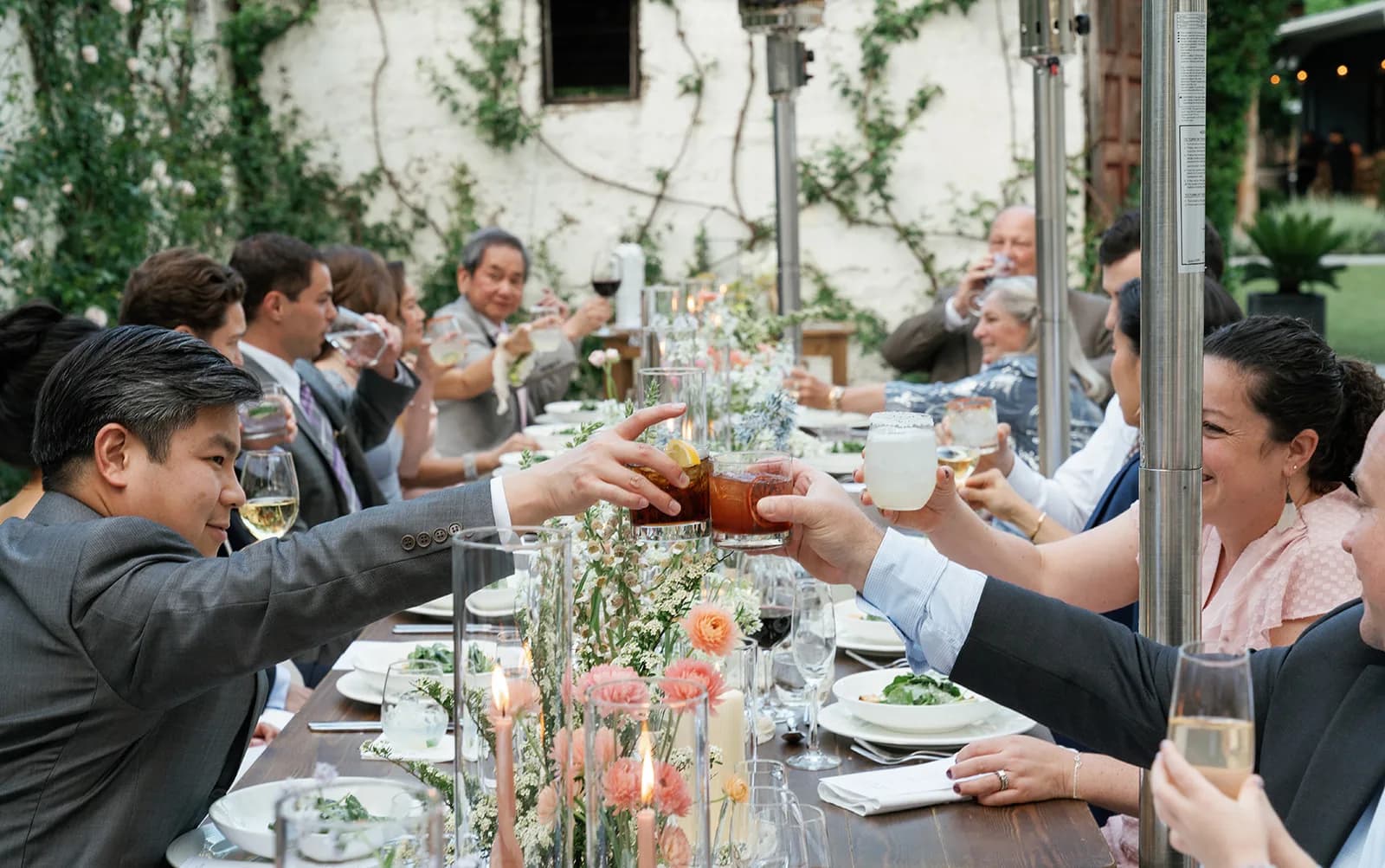 Guests clink glasses across the reception table over lit tapers, coral ranunculus and white lace flower in clustered vases