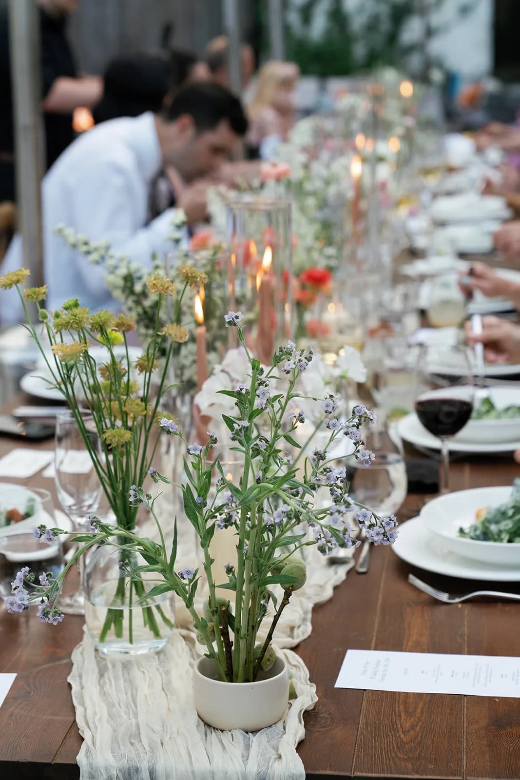 Small yellow blooms and lilac forget-me-not sprays in a glass vase and cream bowl on the runner, candlelight and guests near