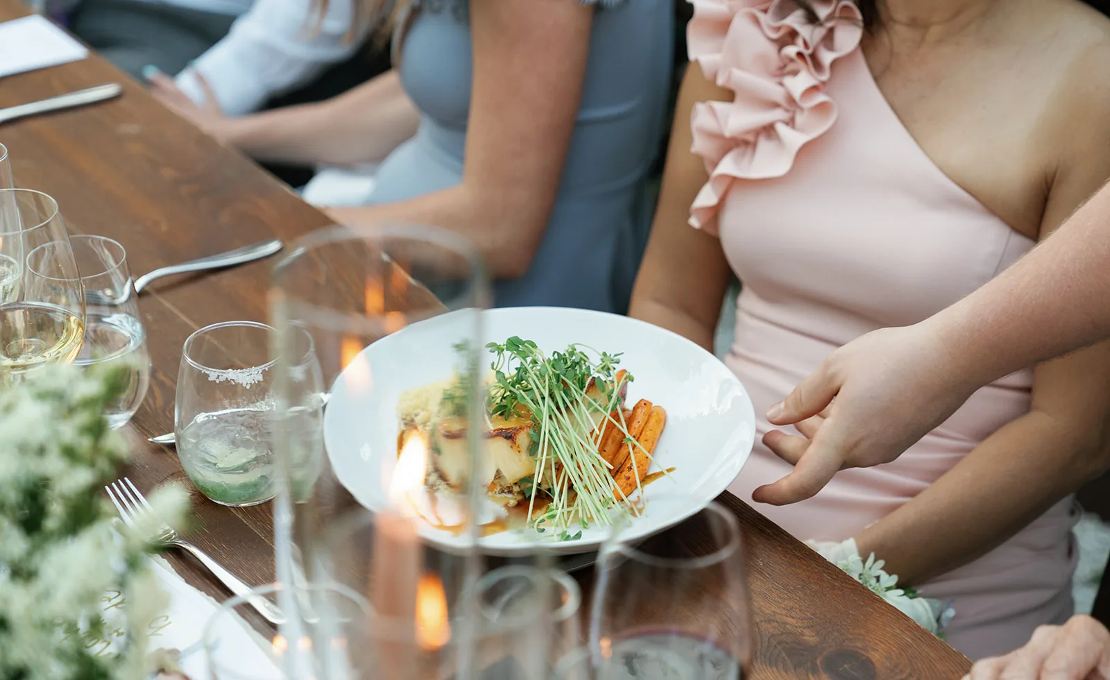 A plated dinner course on the reception table beside a lit taper in glass, white spray blooms soft in the foreground
