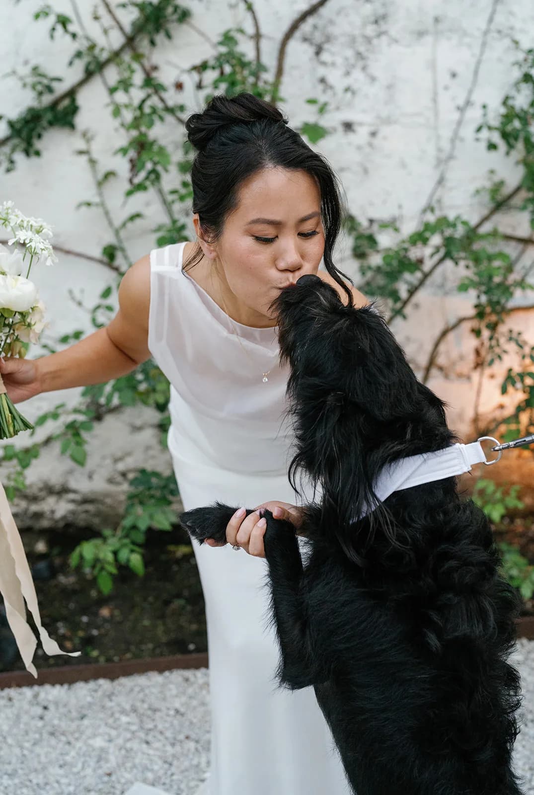 Trisha bends to kiss a black dog standing on its hind legs, holding her white bouquet with silk ribbon in her other hand