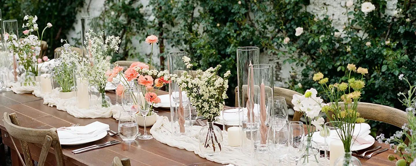 Long wooden table with bud vases of coral poppies, white spirea and sweet pea among taper candles, climbing roses behind
