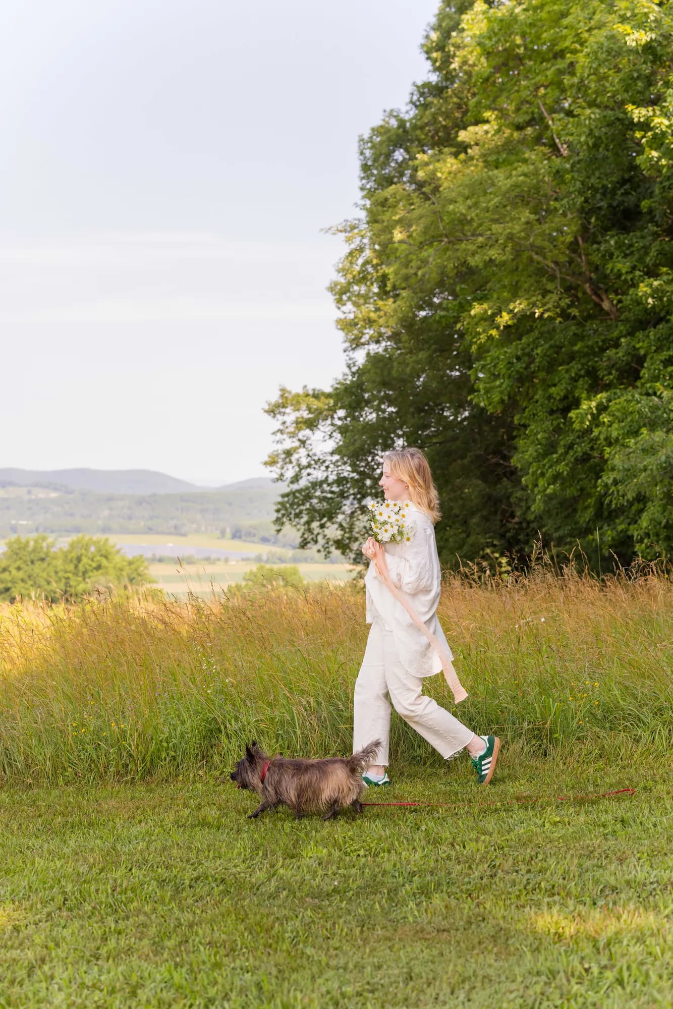 Molly, founder and creative director of Whimsy Flower, walks a hilltop meadow with a bouquet of daisies and her terrier at her heels