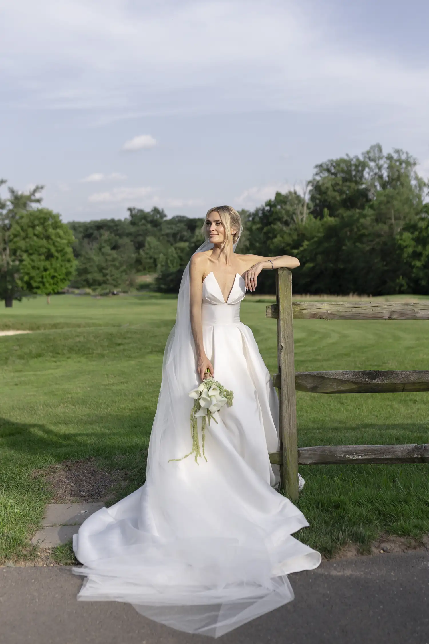 Julia leans on a split-rail fence in a strapless gown and veil, holding a bouquet of white calla lilies and green amaranthus