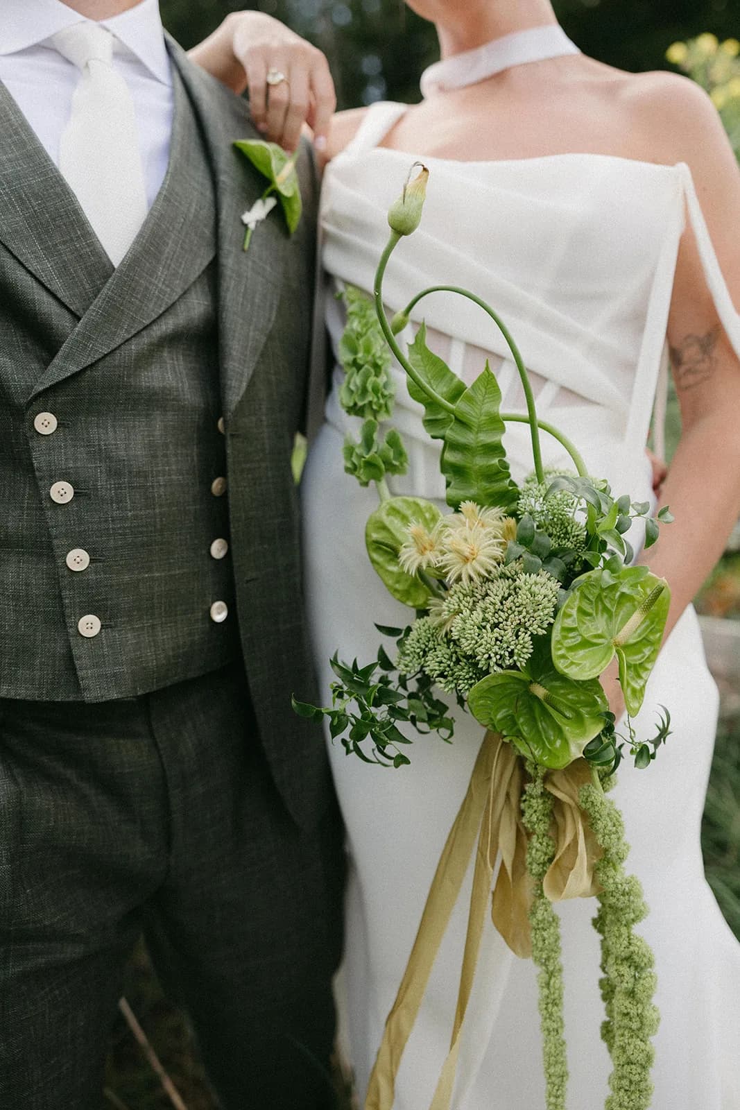 Bride holds a green bouquet of anthurium, bells of Ireland, and trailing amaranthus beside the groom's anthurium boutonniere