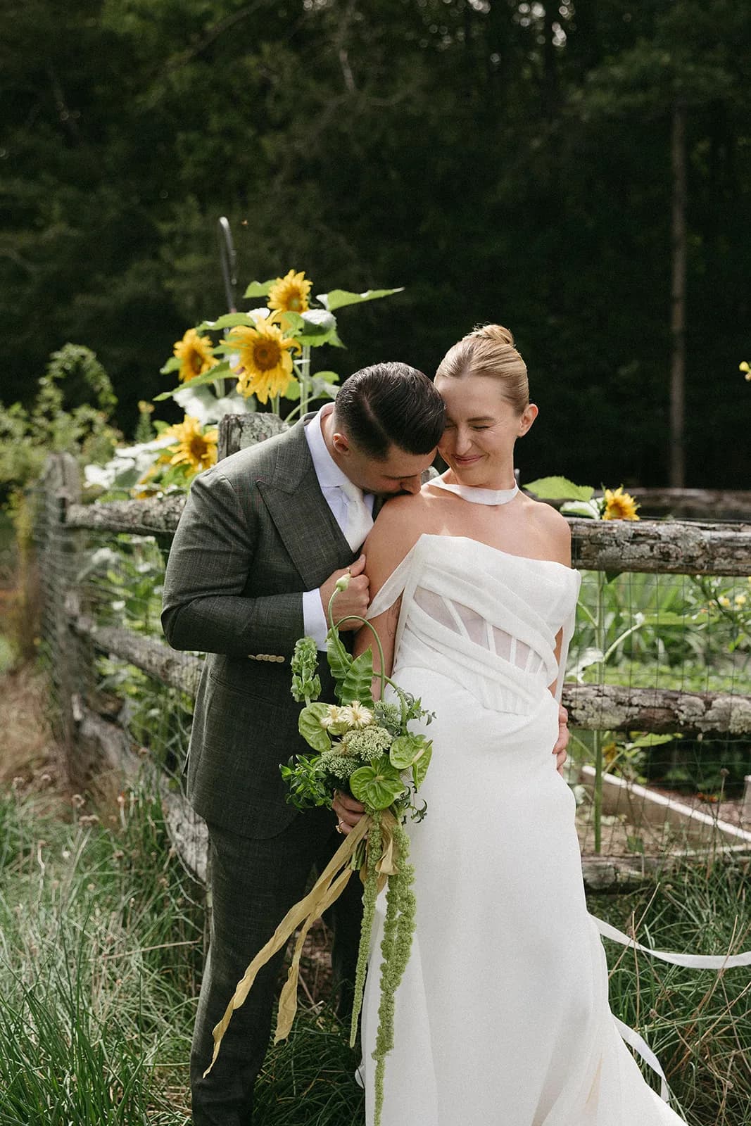 Groom kisses the bride's shoulder at a rustic fence with sunflowers, her bouquet trailing green amaranthus and gold ribbon