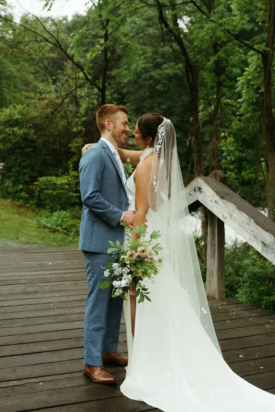 Shayna and Evan embrace on a wooden footbridge, her bouquet of blush scabiosa and pale sweet pea held low at her side