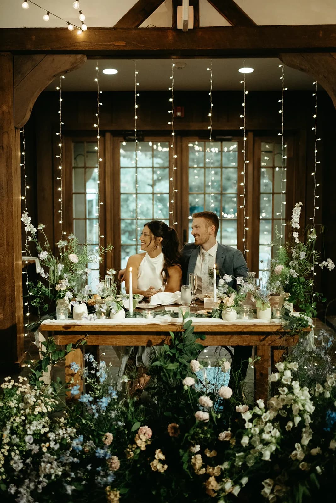 Shayna and Evan at the sweetheart table before a curtain of lights, bud vases and tapers on the runner, blooms massed below