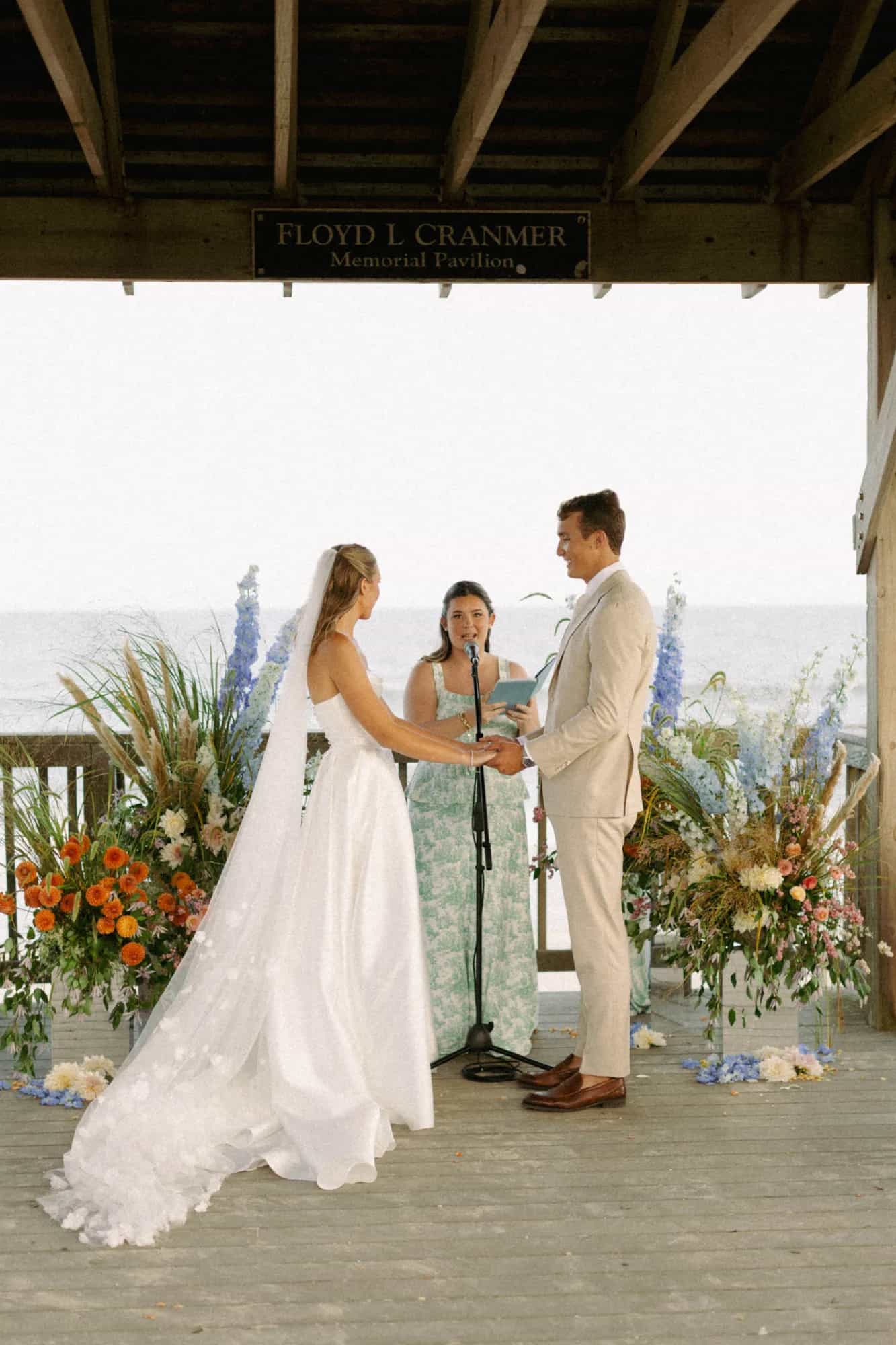 Talea and Erich hold hands at the altar between arrangements on mirror plinths, delphinium and dahlias by the sea