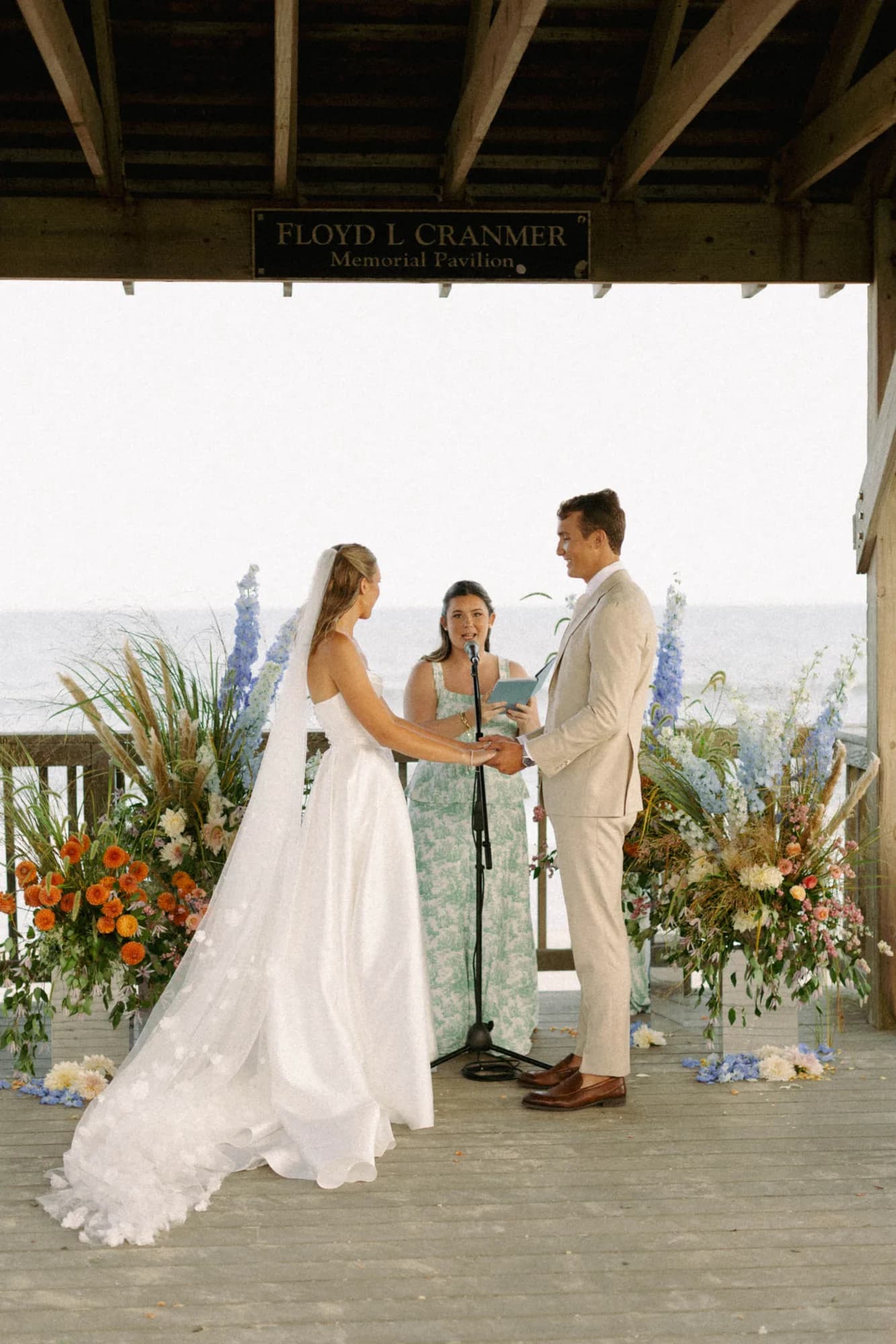 Talea and Erich hold hands at the altar between arrangements on mirror plinths, delphinium and dahlias by the sea