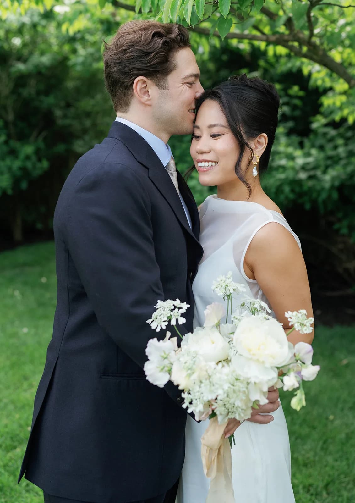 Jim kisses Trisha's temple as she holds her bouquet of white peonies, sweet pea and lace flower tied with silk ribbon