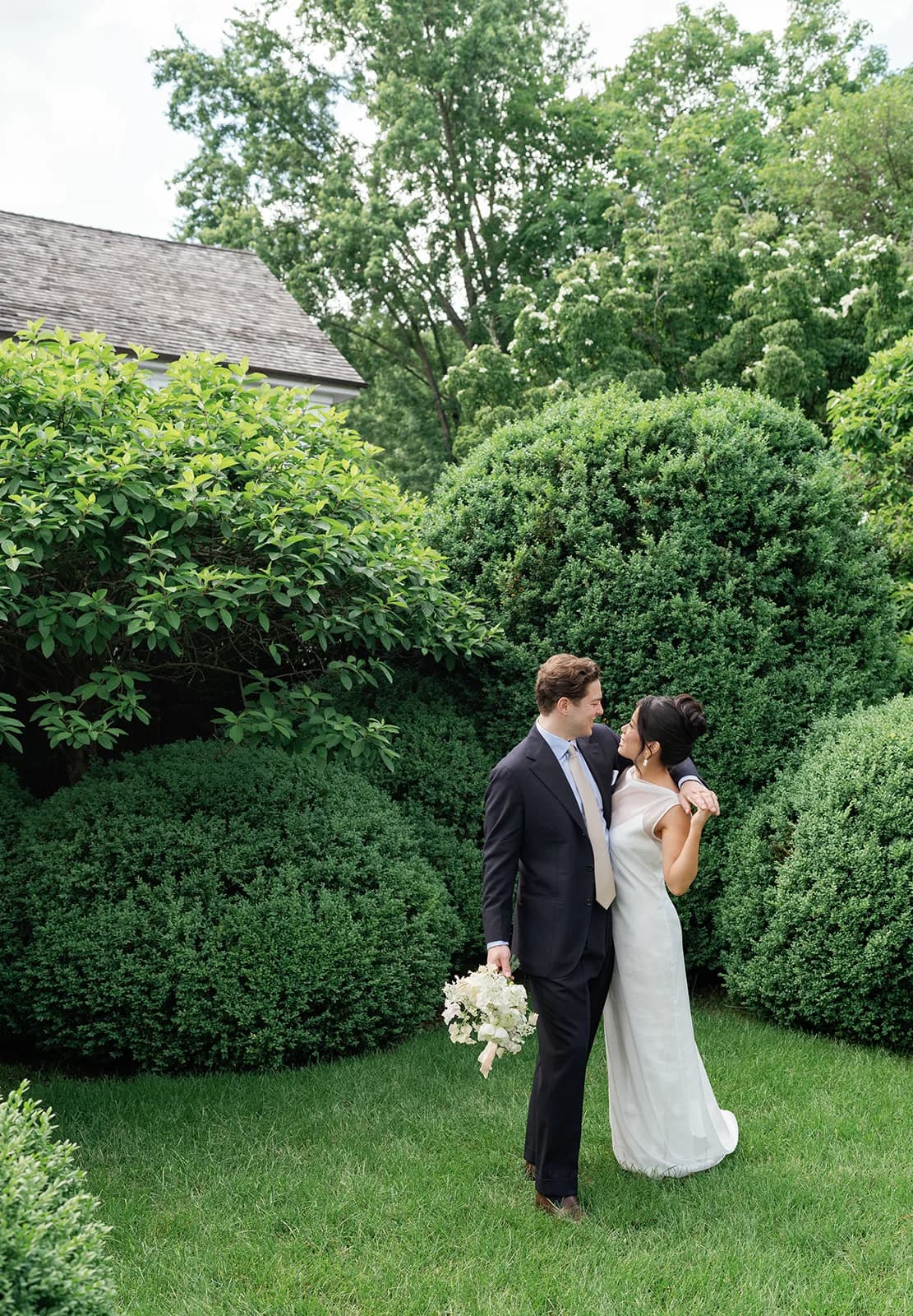 Trisha and Jim walk arm in arm across a lawn between clipped boxwood mounds, Jim carrying her white bouquet at his side