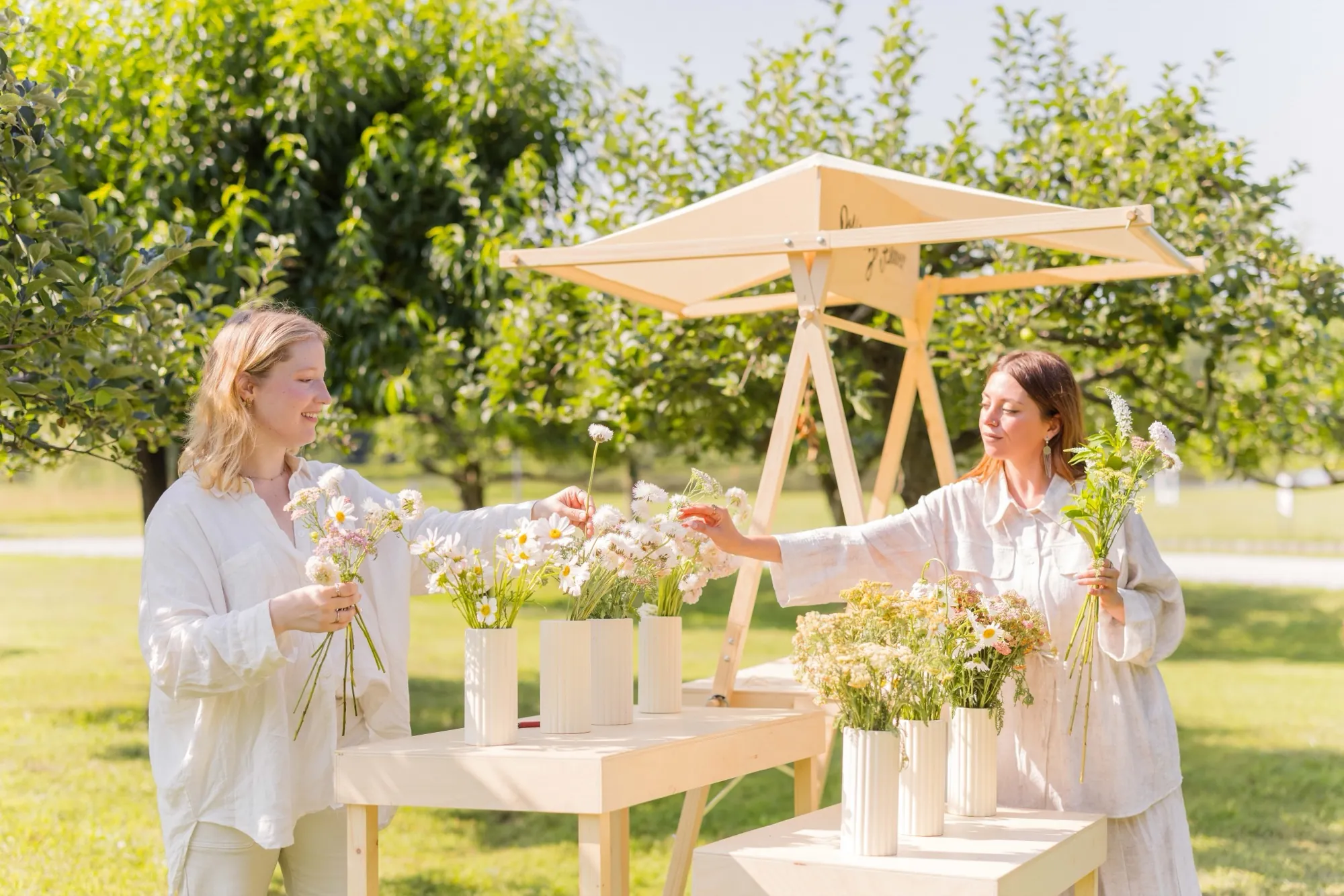 Two women arrange white cosmos, daisies and yarrow in fluted white vessels on a plywood flower stand beneath orchard trees