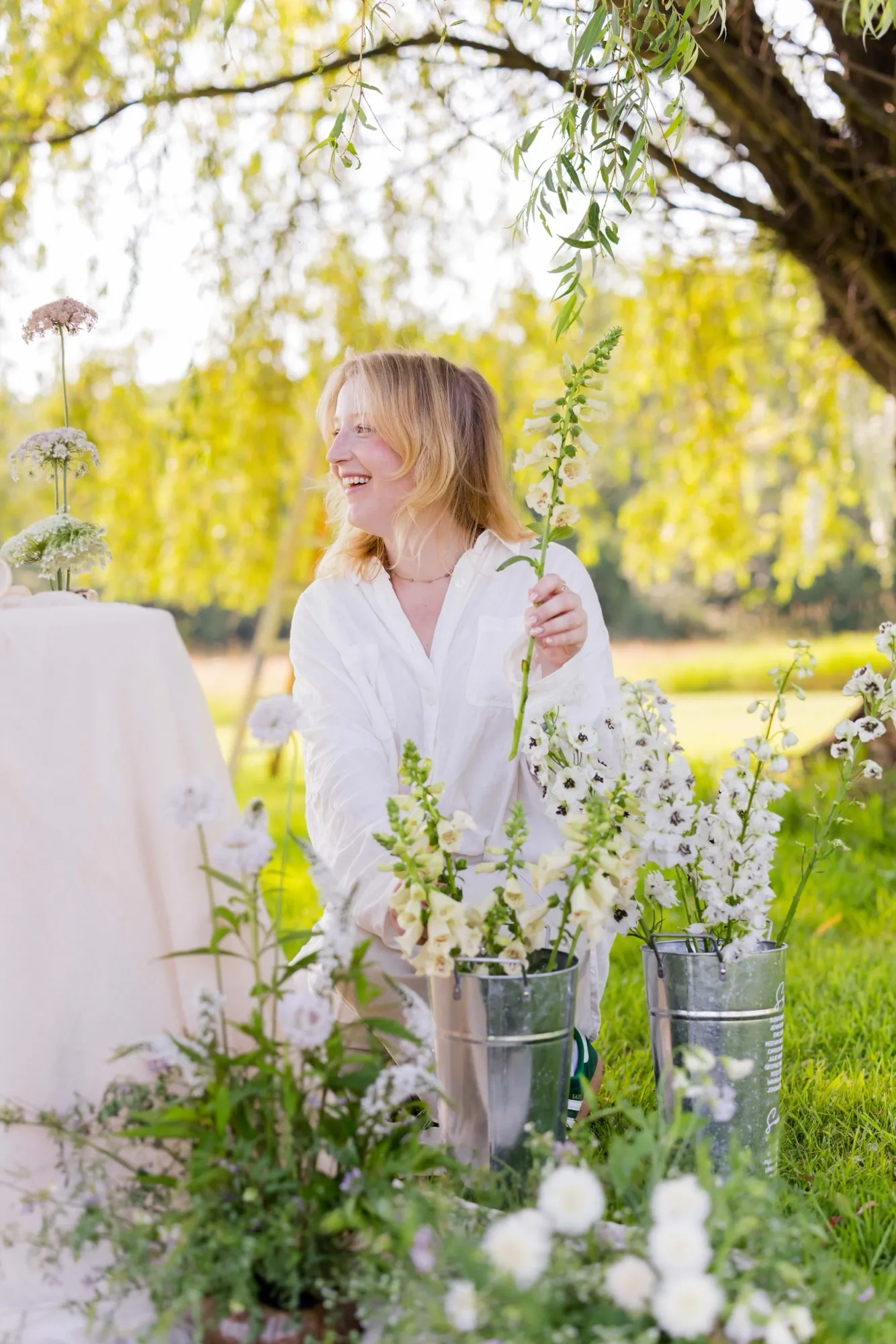 A woman in white lifts a tall stem of cream foxglove beside buckets of white delphinium, sheltered under a willow tree